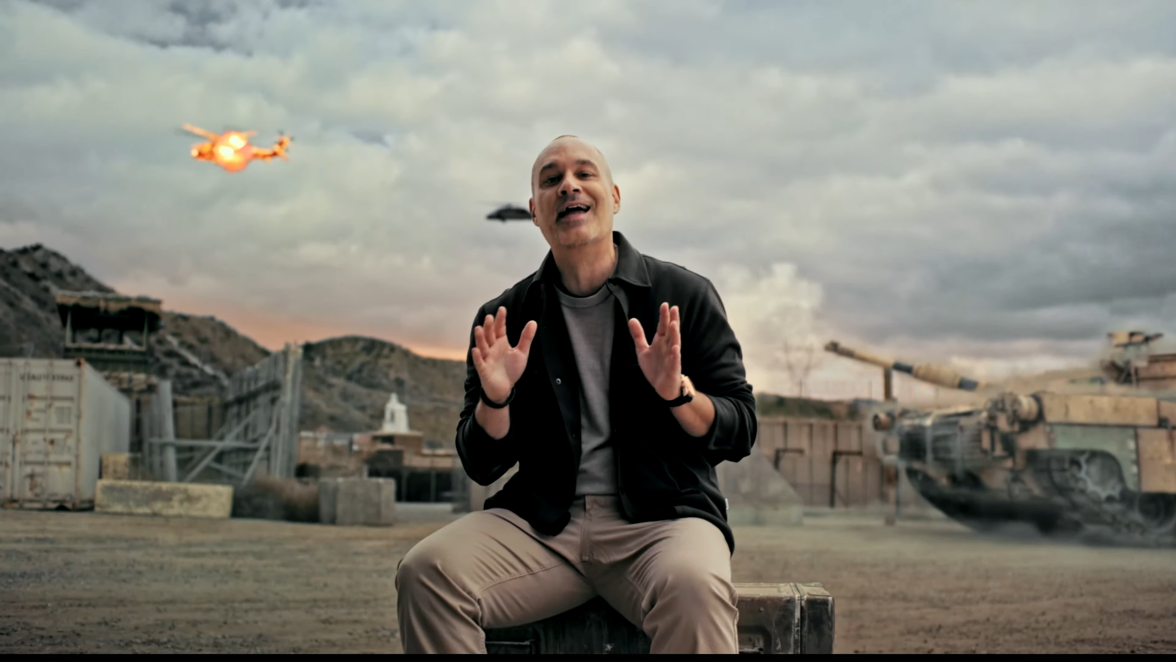 Battlefield executive producer Christian Grass talks to the camera while a helicopter is shot by a tank in the background in the Battlefield 6 multiplayer reveal.