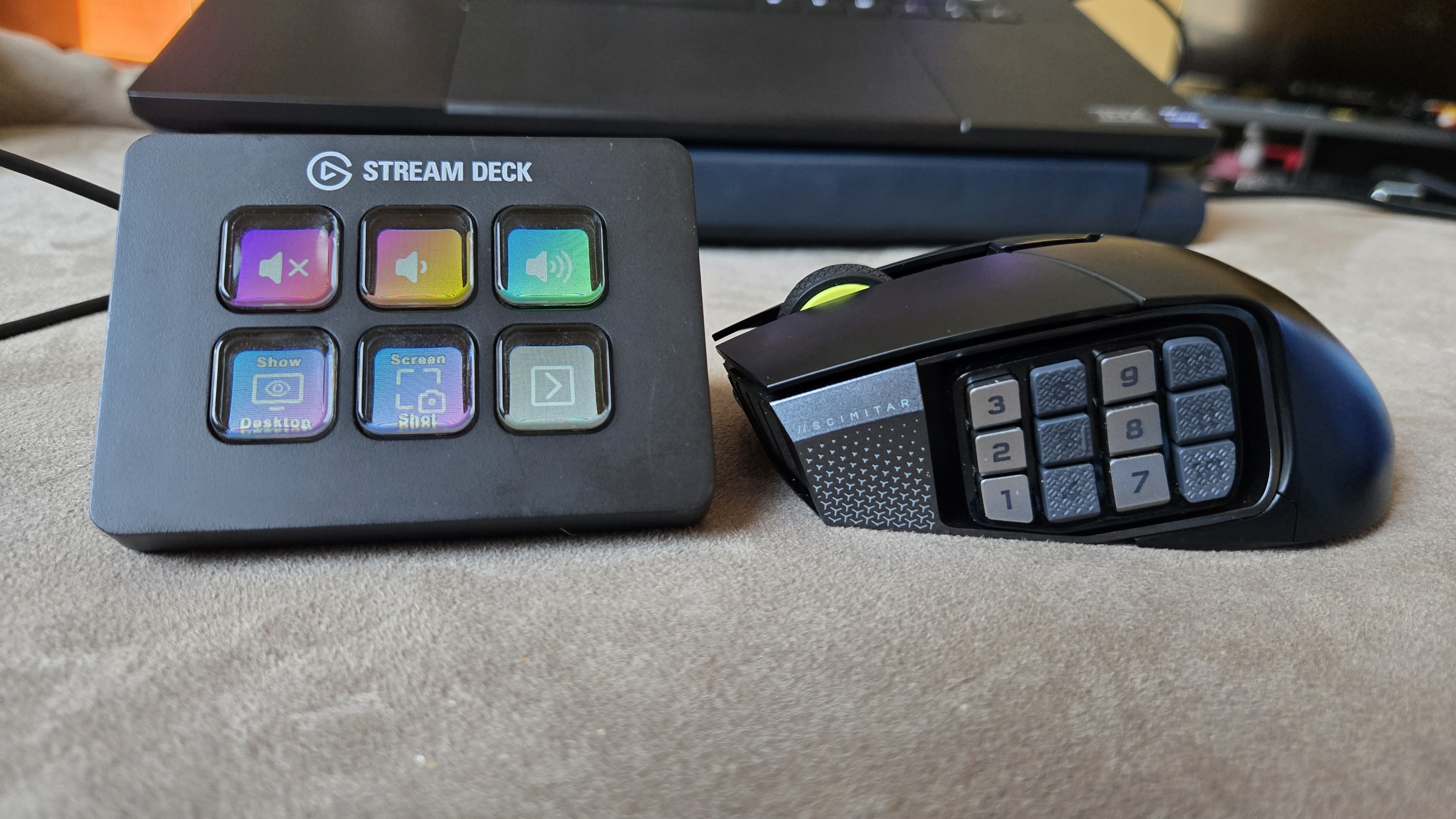 A Corsair Scimitar Elite Wireless SE gaming mouse on a carpet next to a gaming laptop and Stream Deck.