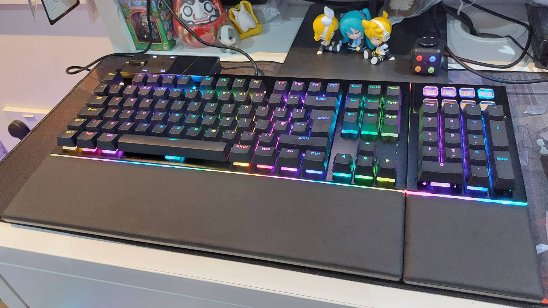 Be Quiet's Dark Mount mechanical keyboard seen on a desk. The modular media dock and numberpad attachments are attached. The two-part magnetic wrist rest is attached. The RGB lights are on, bathing everything in a rainbow-coloured glow.