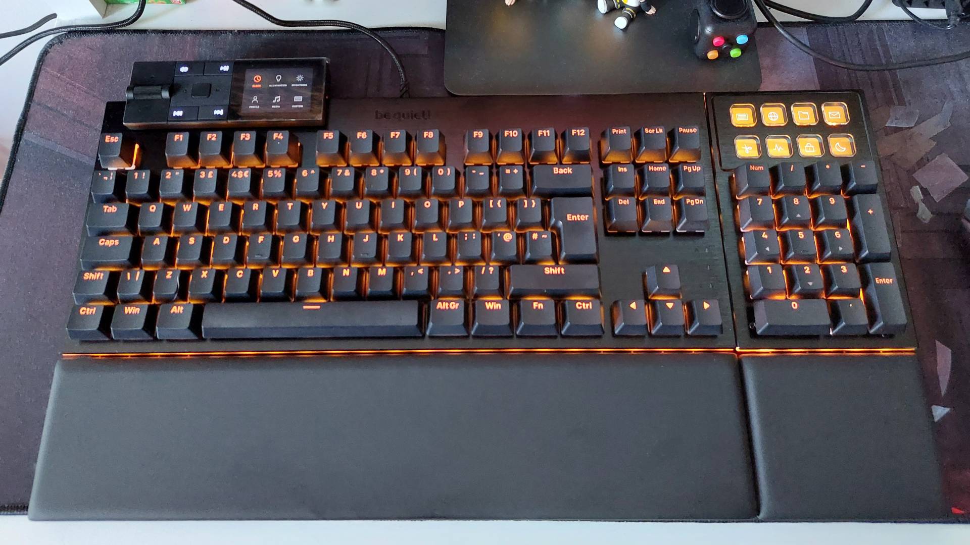 Be Quiet's Dark Mount mechanical keyboard seen on a desk. The modular media dock and numberpad attachments are attached. The two-part magnetic wrist rest is attached. The RGB lights are on, bathing everything in orange light.