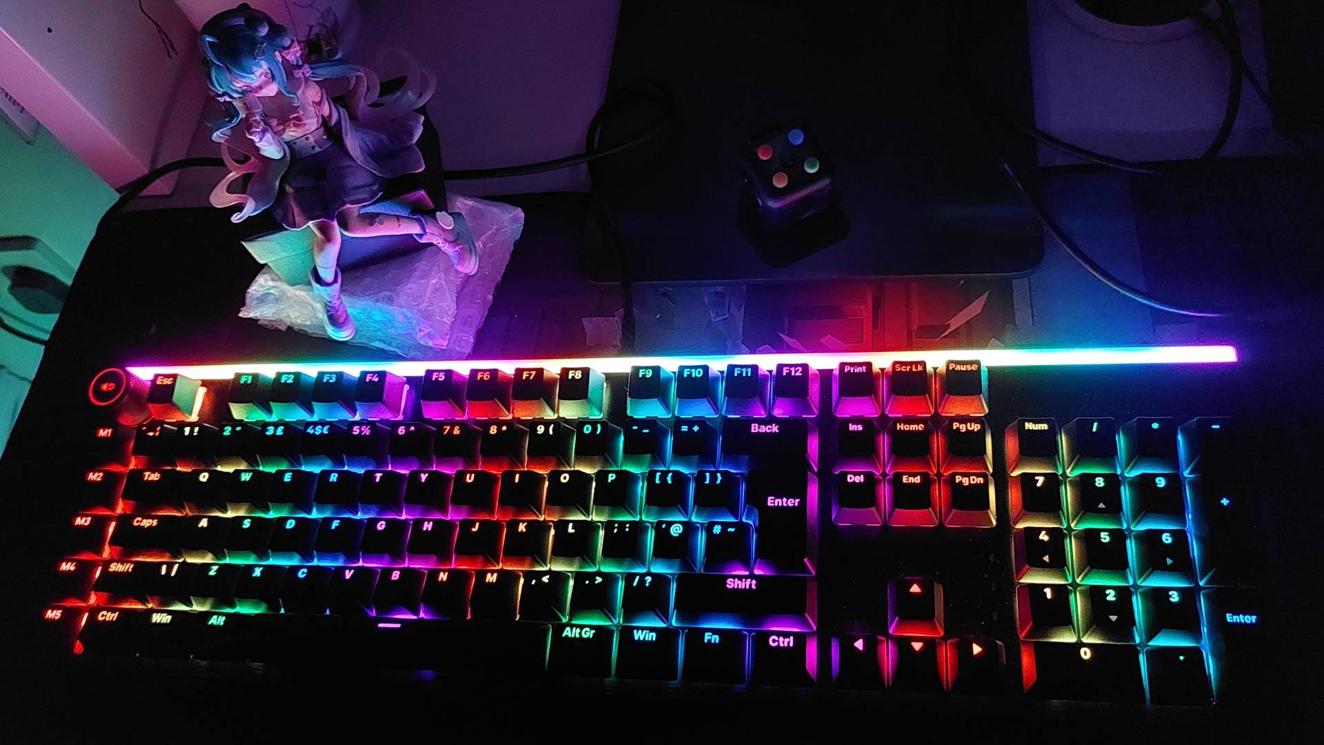 In a darkened room, Be Quiet's Light Mount gaming mechanical keyboard can be seen with its RGB lights on. The RGB lights are one of the only light sources in the dark room, illuminating a nearby Love Sailor Miku Hatsune anime figurine, and a fidget cube.