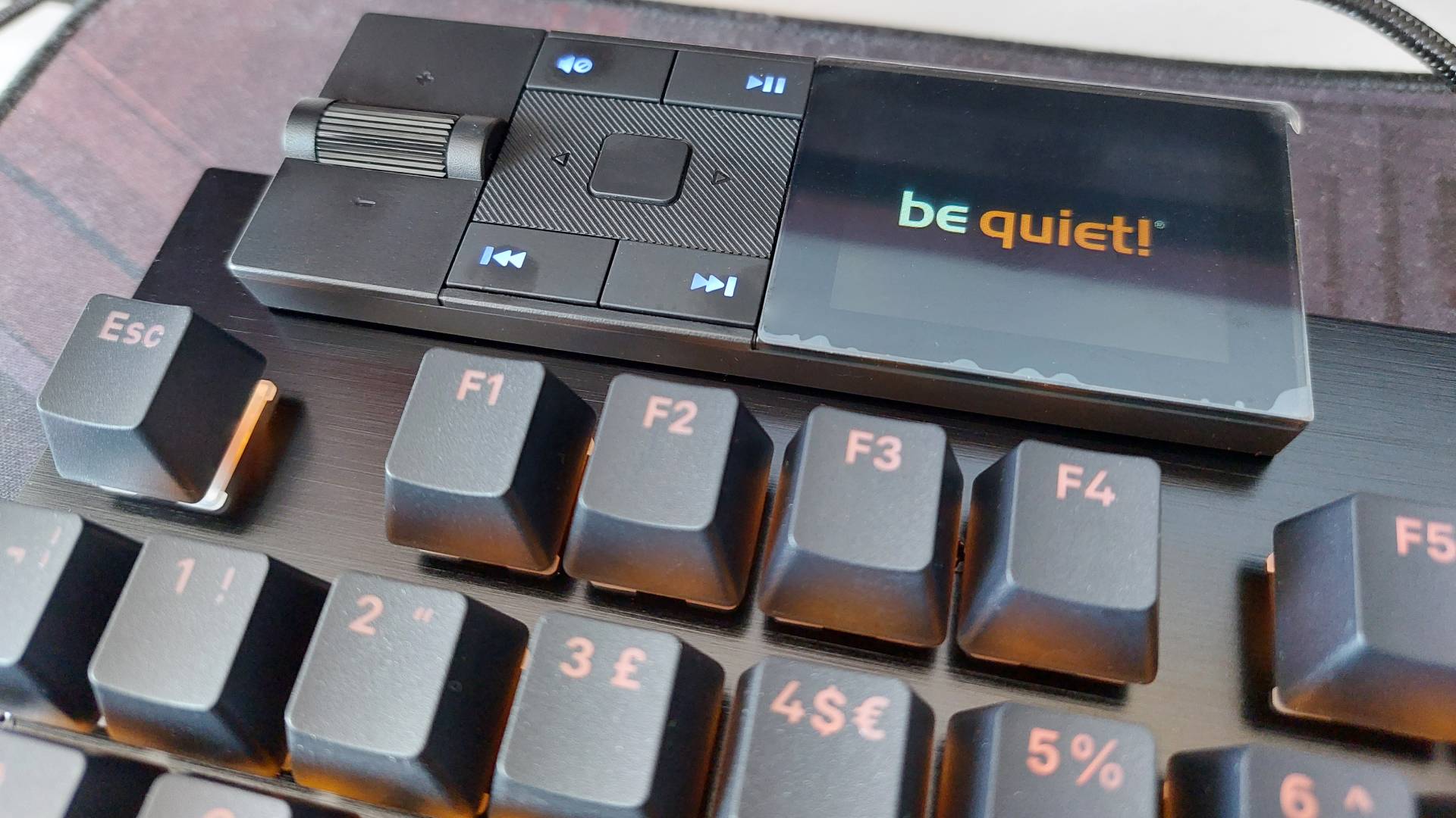 A close up of the Dark Mount mechanical keyboard's modular media dock attachment. It's switched on, displaying the be quiet! logo on its inbuilt screen. The keyboard's RGB light are also on, bathing everything in an orange light.