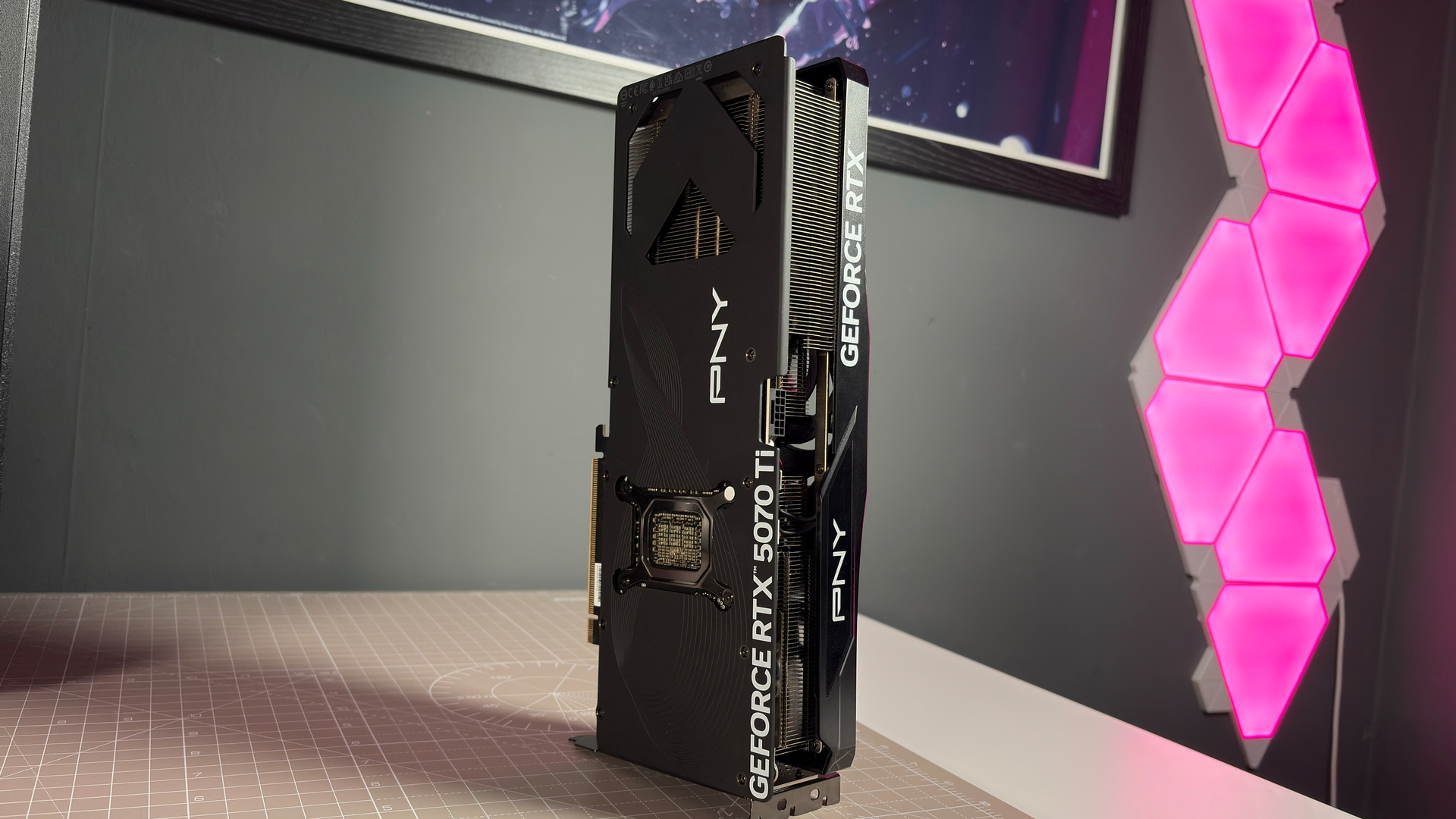 A PNY RTX 5070 Ti OC graphics card on a desk with a desk mat and pink light.
