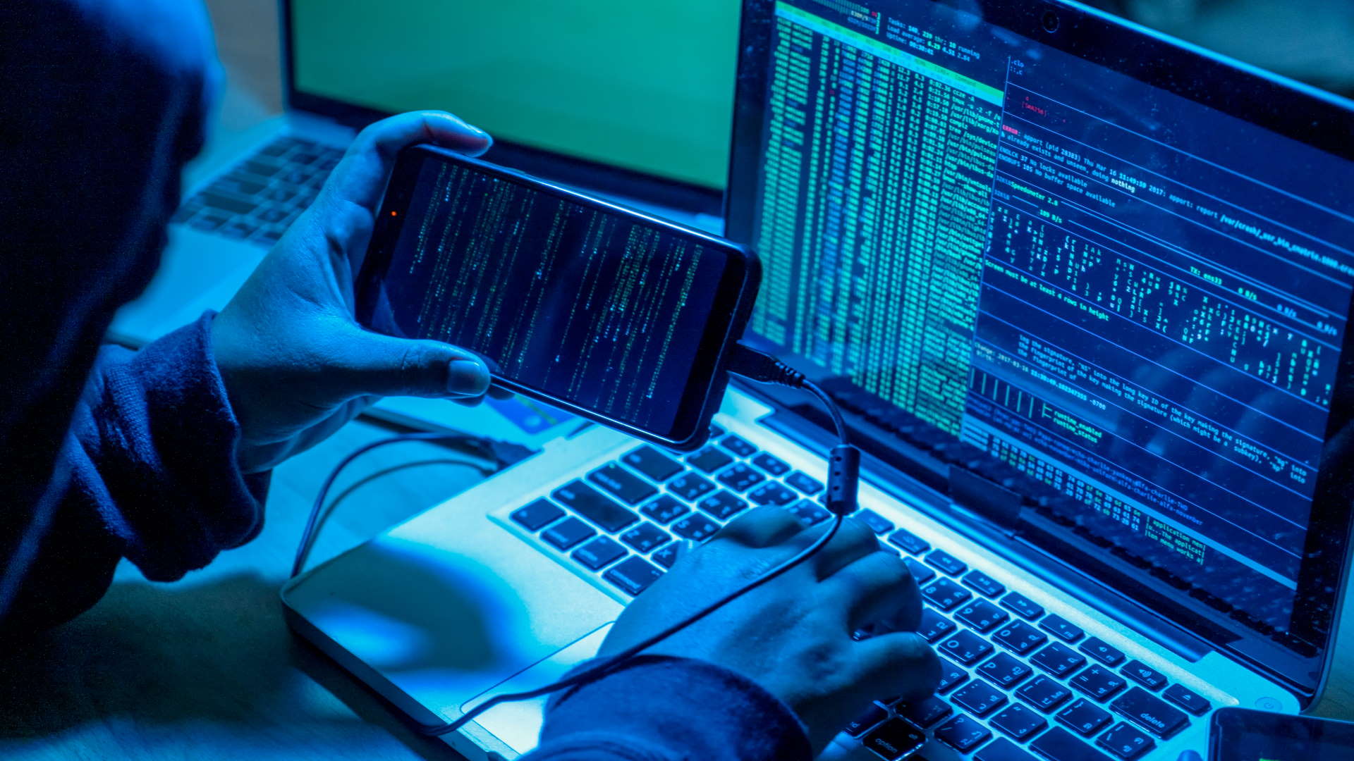 A stock photo of a hacker with computers in dark room. The devices are displaying computer code on the screens.