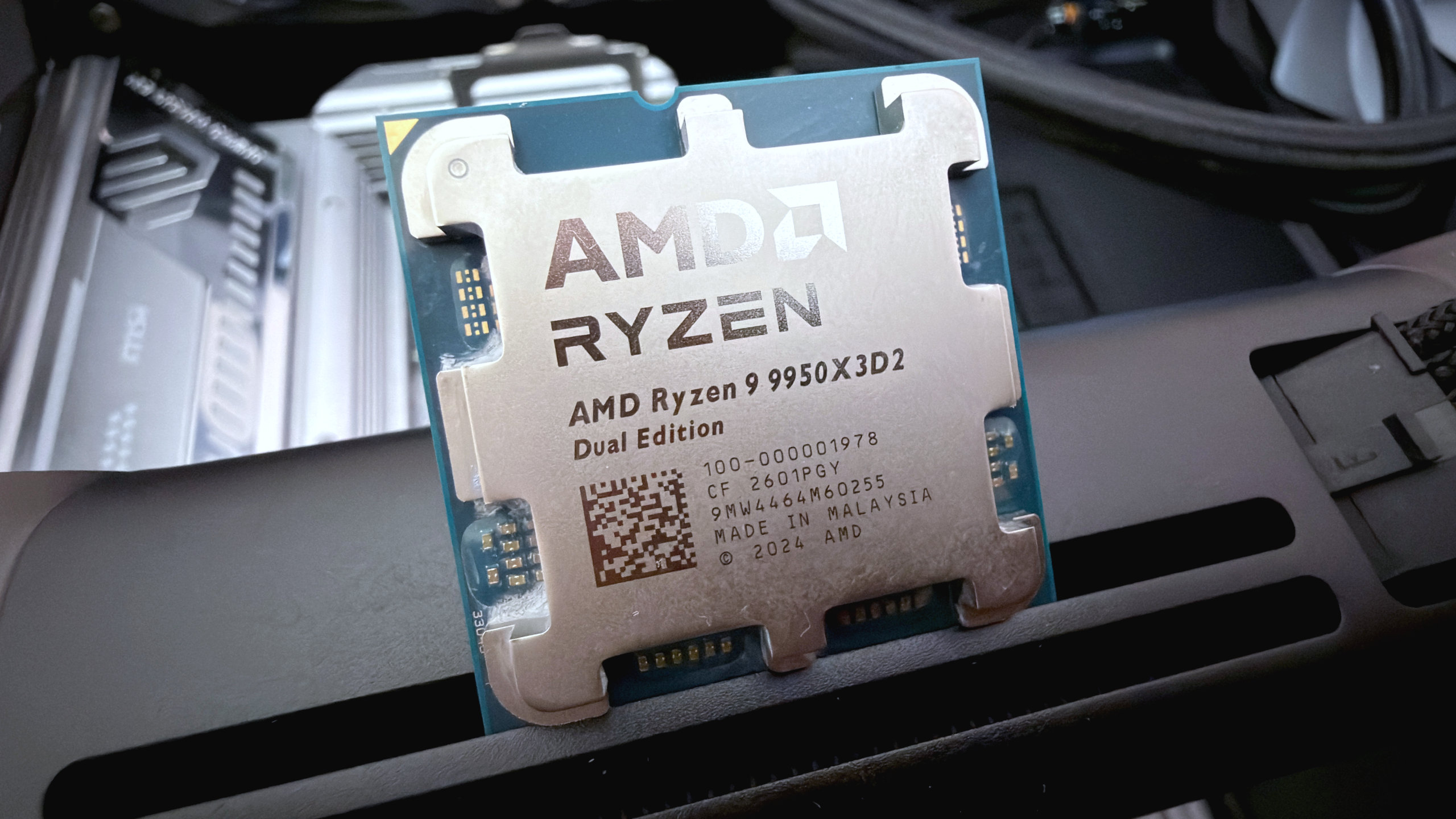 A photo of an AMD Ryzen 9 9950X3D2 Dual Edition processor, with PC components in the background