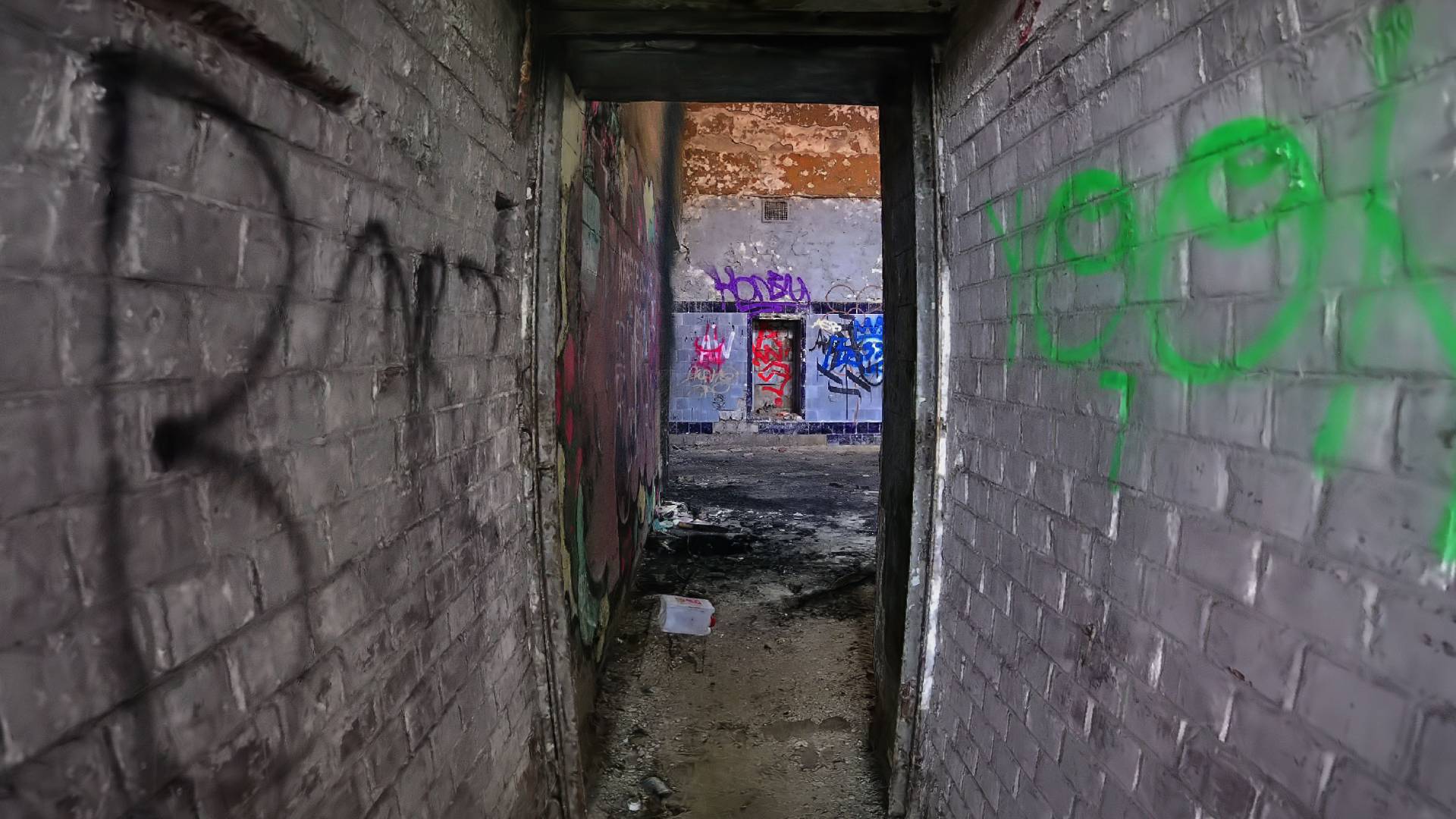 A Gaussian splat scene captured by scan artist Christoph Schindelar. This is a narrow corridor in an abandoned factory. The walls are white-painted brick, with graffitti tags scrawled over the top.