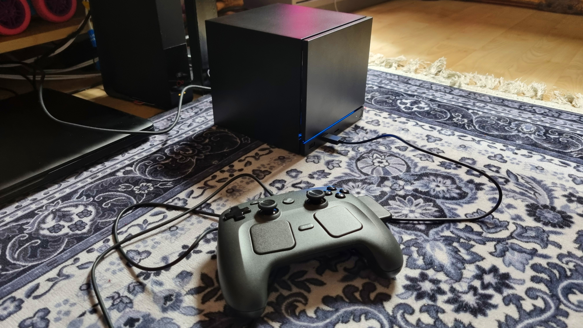 A Steam Machine in front of a TV, with the Steam Controller resting against it and a pink neon sign in the background