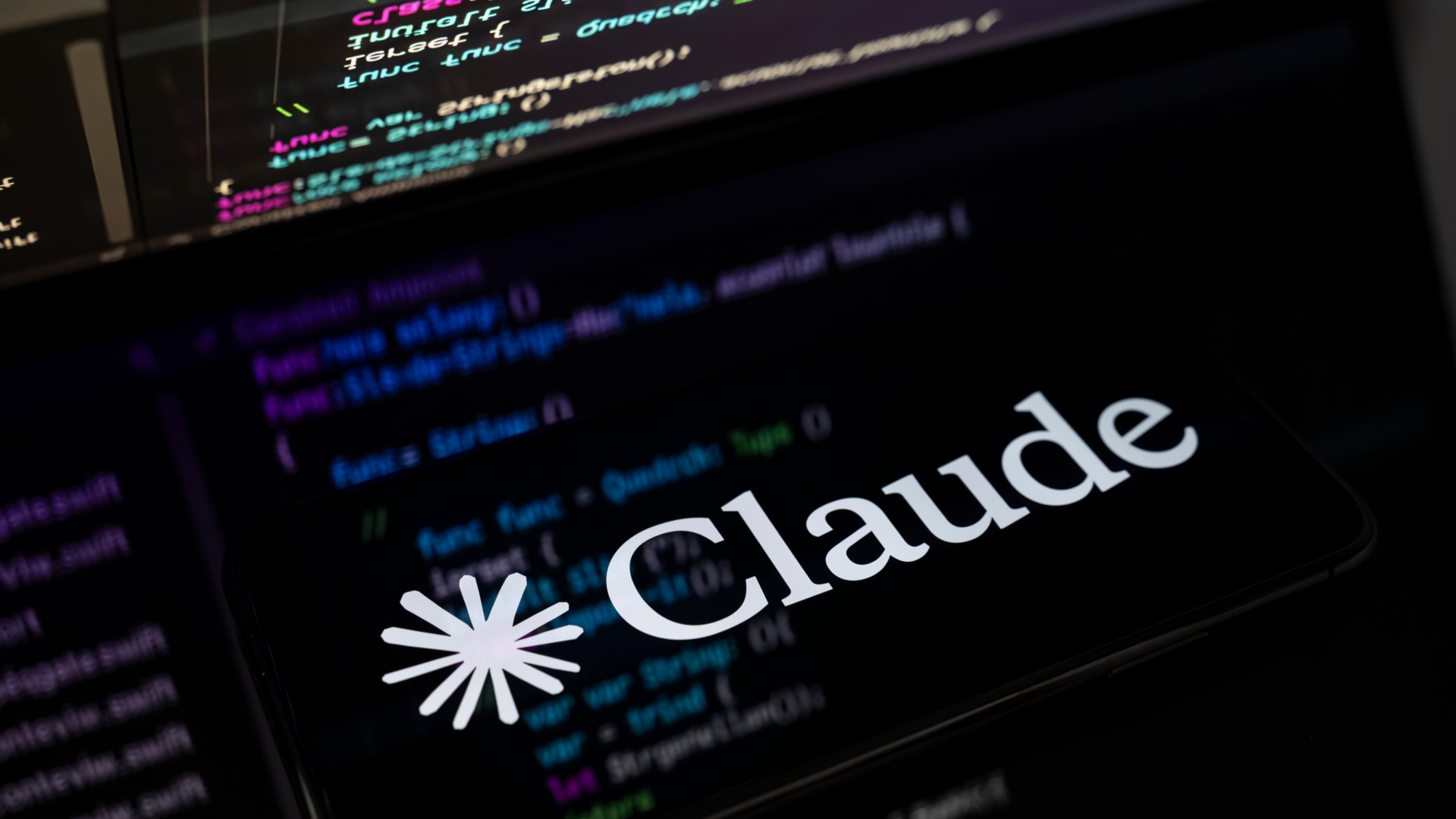 An image showing the Claude AI logo displayed on the screen of a smartphone placed on a reflective surface onto which lines of computer code are projected.